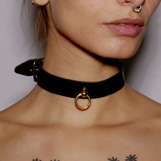 Choker fin en cuir noir surmonté d'un anneau en acier doré. Il se porte comme bijou ou accessoire de jeu de soumission